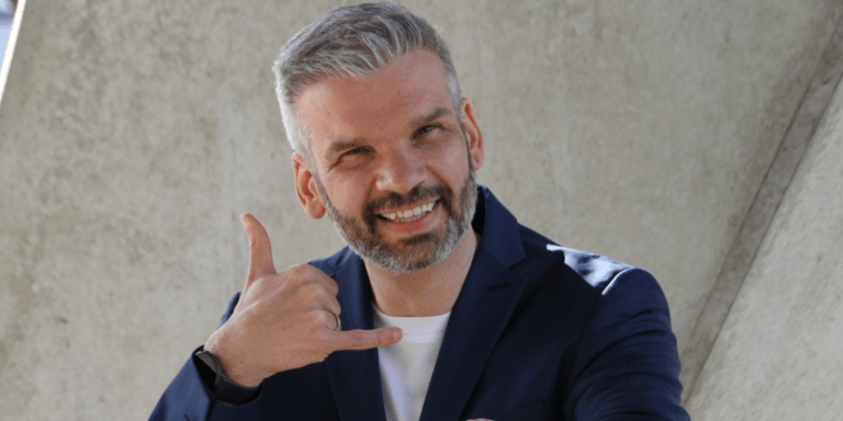 Mittelaufnahme von Business Coach Florian Fassnacht im marineblauen Sakko und Vollbart, der lächelnd die „Ruf mich an“-Geste macht.